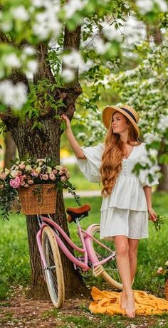 a woman standing next to a pink bike under a tree with flowers on the basket