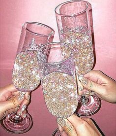 three people holding up wine glasses filled with liquid and sparkles on pink wall background