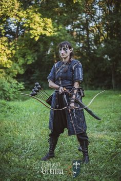 a woman dressed in black holding two bows and arrows with trees in the back ground