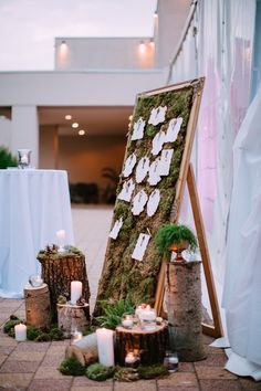 an easel with moss and candles on the ground in front of a white table cloth