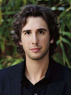 a close up of a person wearing a black shirt and blazer with trees in the background