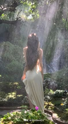 a woman standing in front of a waterfall with her back to the camera and sunlight streaming down