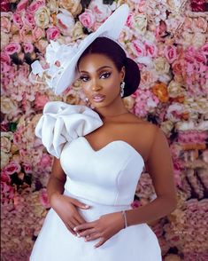 a woman in a white dress and hat posing for the camera with flowers behind her