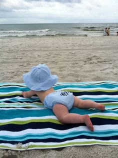 a baby laying on top of a blue and green blanket next to the ocean with people in the background