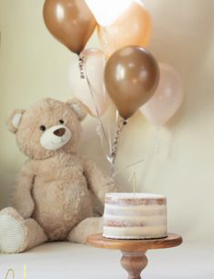 a teddy bear sitting next to a cake with balloons in the air and a candle on top