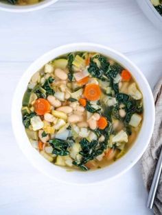 two bowls filled with soup and vegetables on top of a white table next to silver spoons