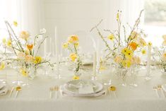 the table is set with yellow and white flowers in vases, candles, and plates