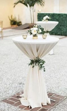 a white table topped with flowers and greenery on top of a stone floor next to two vases