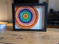 a multicolored paper doily in a black frame on a wooden table