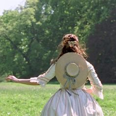 a woman in a dress and hat is walking through the grass with her arms outstretched