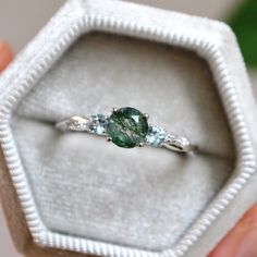 a close up of a person holding an engagement ring in a white box with green and blue stones