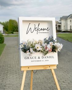 a welcome sign with flowers on it sitting in front of a brick road and house