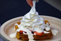 a waffle topped with whipped cream and strawberries on a paper plate, being drizzled with syrup