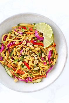 a white bowl filled with noodles and vegetables next to a lime wedge on the side