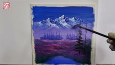 a person is holding a paintbrush in front of a painting with mountains and trees on it