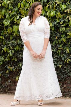 a woman wearing a white dress standing in front of a bush with her hands on her hips