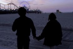 two people holding hands while walking on the beach at dusk with ferris wheel in background