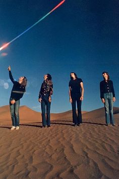 four people standing on top of a sand dune flying a kite in the blue sky