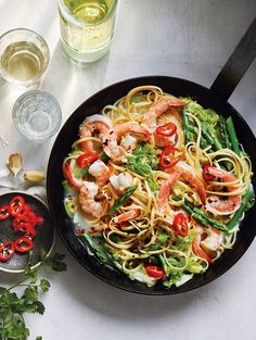 a pan filled with pasta and shrimp on top of a table next to wine glasses