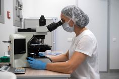 a woman in white shirt and blue gloves looking through a microscope at something on the table