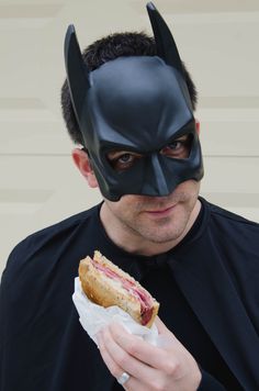 a man wearing a batman mask and eating a sandwich with meat in his hand while looking at the camera