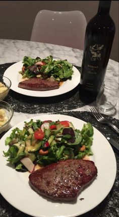 two white plates topped with steak and salad next to a bottle of wine on a table