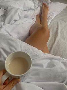 a woman laying in bed with her feet on the pillow and holding a cup of coffee