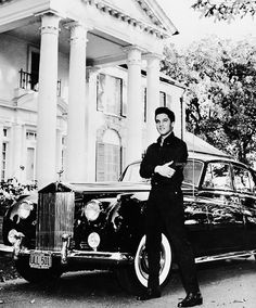 a black and white photo of a man standing next to a classic car in front of a house