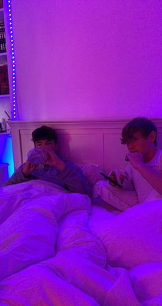 two young boys laying in bed with purple lights on the headboard and one holding a camera