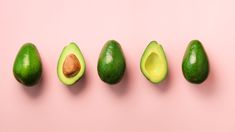 five avocados lined up in a row on a pink background with one cut in half