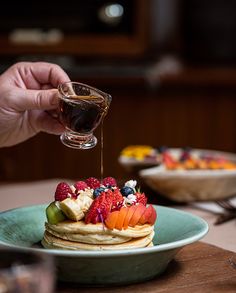 a person pouring syrup on top of a stack of pancakes covered in berries and kiwis