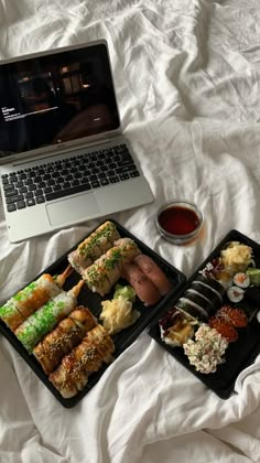 two trays of sushi next to an open laptop on a bed with white sheets