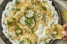 a person holding a cracker in front of a bowl of dip with cucumbers and dill