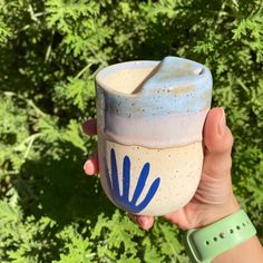 a hand holding a cup with blue paint on it in front of some green plants