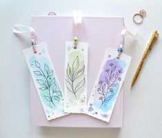 three bookmarks with watercolor designs on them next to a pen and pencils