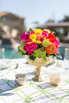 colorful flowers in a gold vase on a table next to some glasses and silverware