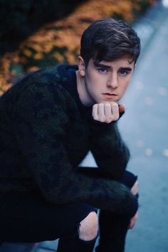 a young man is sitting on the curb with his hand under his chin and looking at the camera