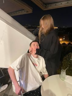 two people standing on top of a roof next to a table and chair with drinks