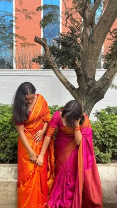 two women in orange and pink sari standing next to each other near a tree