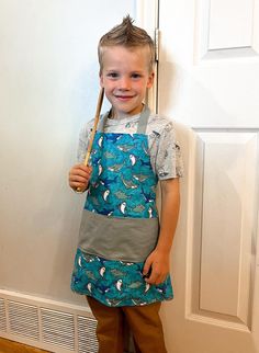 a young boy standing in front of a door holding a wooden stick and wearing an apron