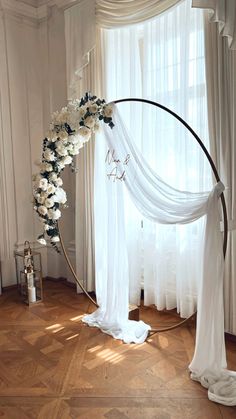 a white wedding arch with flowers on the top and curtains in the back, sitting next to a window