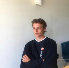 a young man with his arms crossed standing in front of a desk and chair, looking at the camera