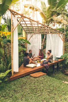 three people sitting on a wooden bench in the middle of a jungle area with white curtains