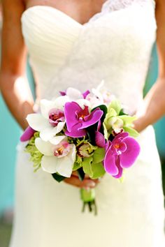 a bride holding a bouquet of purple and white flowers on her wedding day in front of a turquoise wall