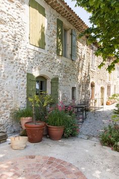 an outdoor patio with potted plants and tables in front of a stone building that has green shutters