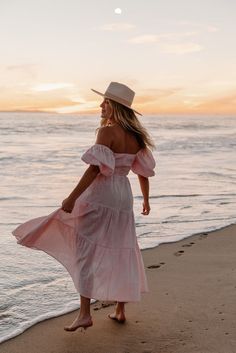 a woman in a pink dress and hat walks along the beach at sunset with her back to the camera