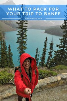 a woman in red jacket standing on top of a mountain next to trees and water