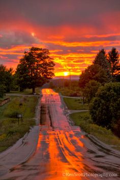 the sun is setting over an empty road with puddles on it and trees in the background