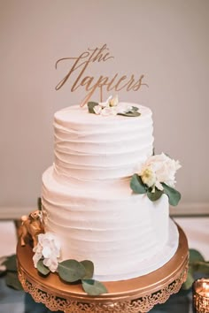 a white wedding cake sitting on top of a gold platter with flowers and greenery