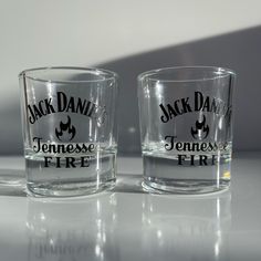 two shot glasses sitting next to each other on top of a white table with the words jack daniels and tennessee fire printed on them